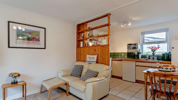 Un salon comprenant un canapé, un banc en bois et une table à manger avec des chaises. Il y a également un coin cuisine avec un évier, une cuisinière et des placards. Un tableau encadré est accroché au mur.