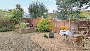 Un jardin avec un chemin de gravier, une clôture en bois, des plantes en pot, une table avec des chaises et un drapeau.