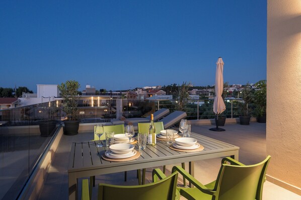 A rooftop dining area with a table set for two, potted plants, and a city view at dusk. - Altana Sunset Living (Agia Marina)