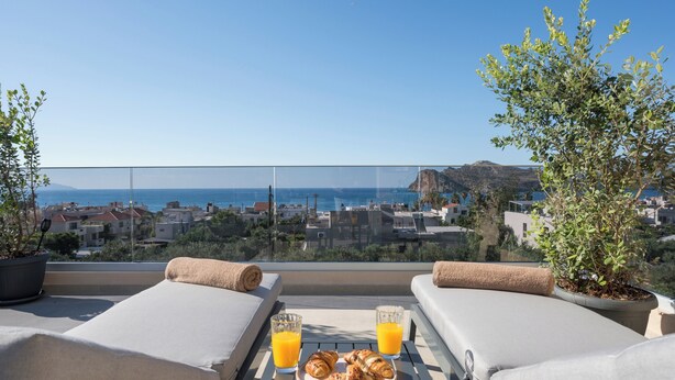 Een balkon met twee loungestoelen, een tafel met croissants en sinaasappelsap, en uitzicht op zee en gebouwen.