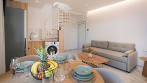 Een moderne woonkamer met een bank, een eettafel met fruit en glazen, een keuken met een wasmachine en een spiltrap.