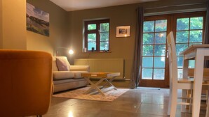Ein Wohnzimmer mit Sofa, Sessel, Tisch und Heizkörper. Es gibt Fenster mit Vorhängen und ein Bild an der Wand.