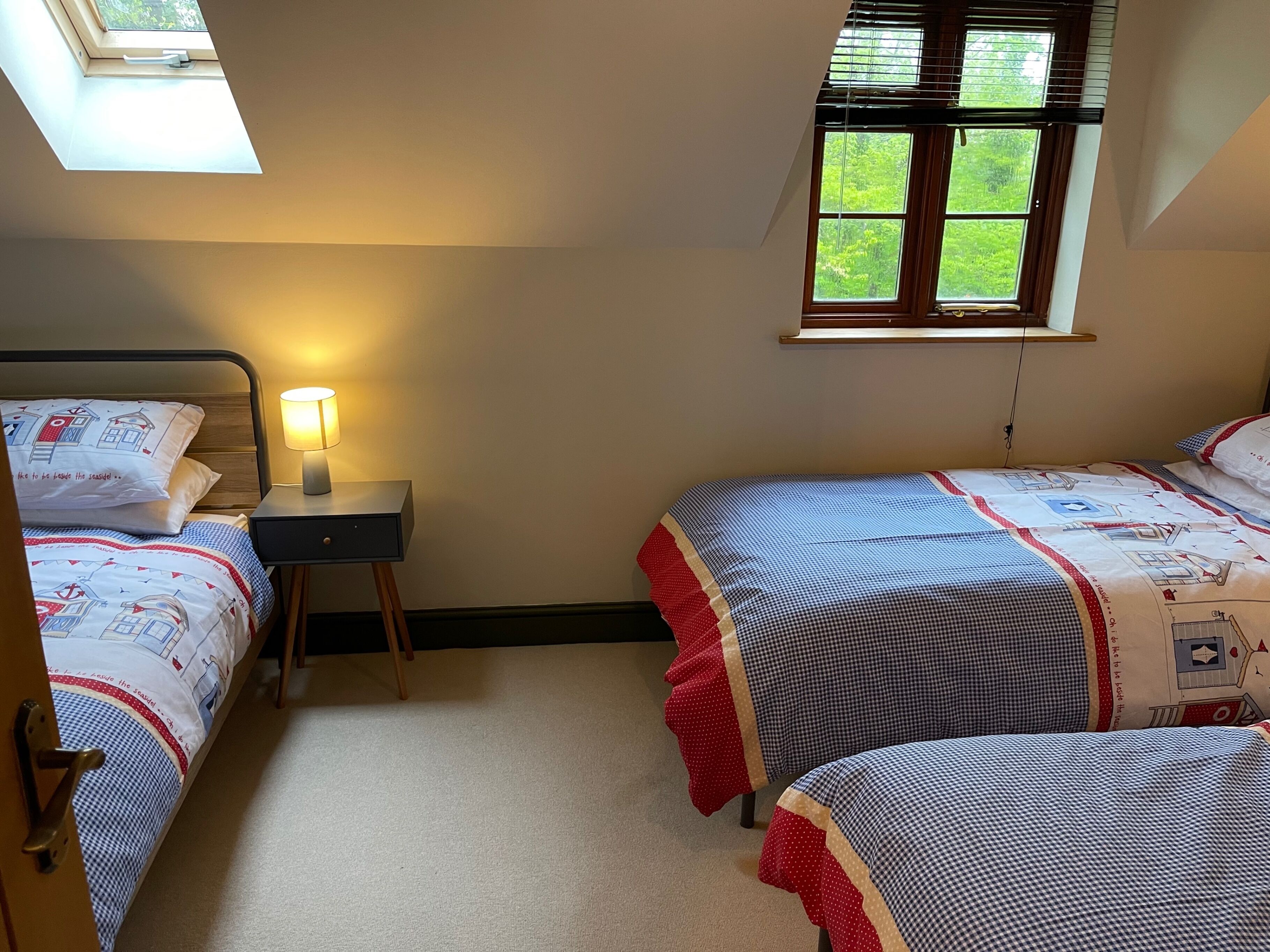 A bedroom with two beds, a bedside table with a lamp, a window with blinds, and a view of greenery outside.