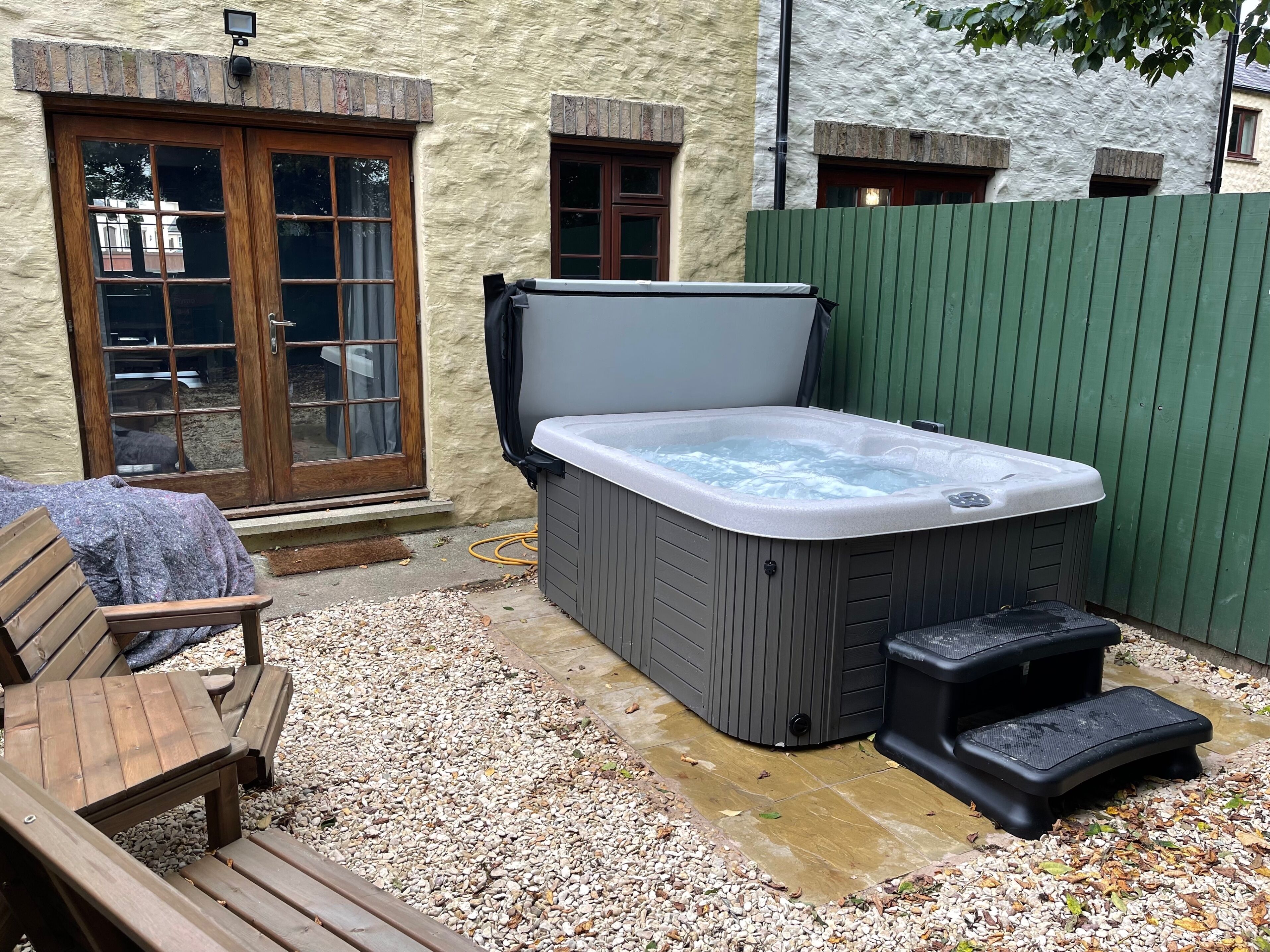 A hot tub, wooden deck, and a bench in a backyard with a stone wall and a green fence.