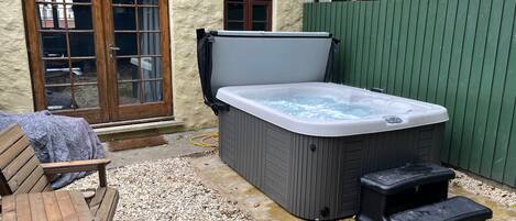A hot tub, wooden deck, and a bench in a backyard with a stone wall and a green fence.