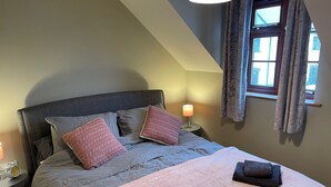Ein Schlafzimmer mit Bett, Kissen, einer Nachttischlampe, einem Fenster mit Vorhängen und einem Handtuch auf dem Bett.