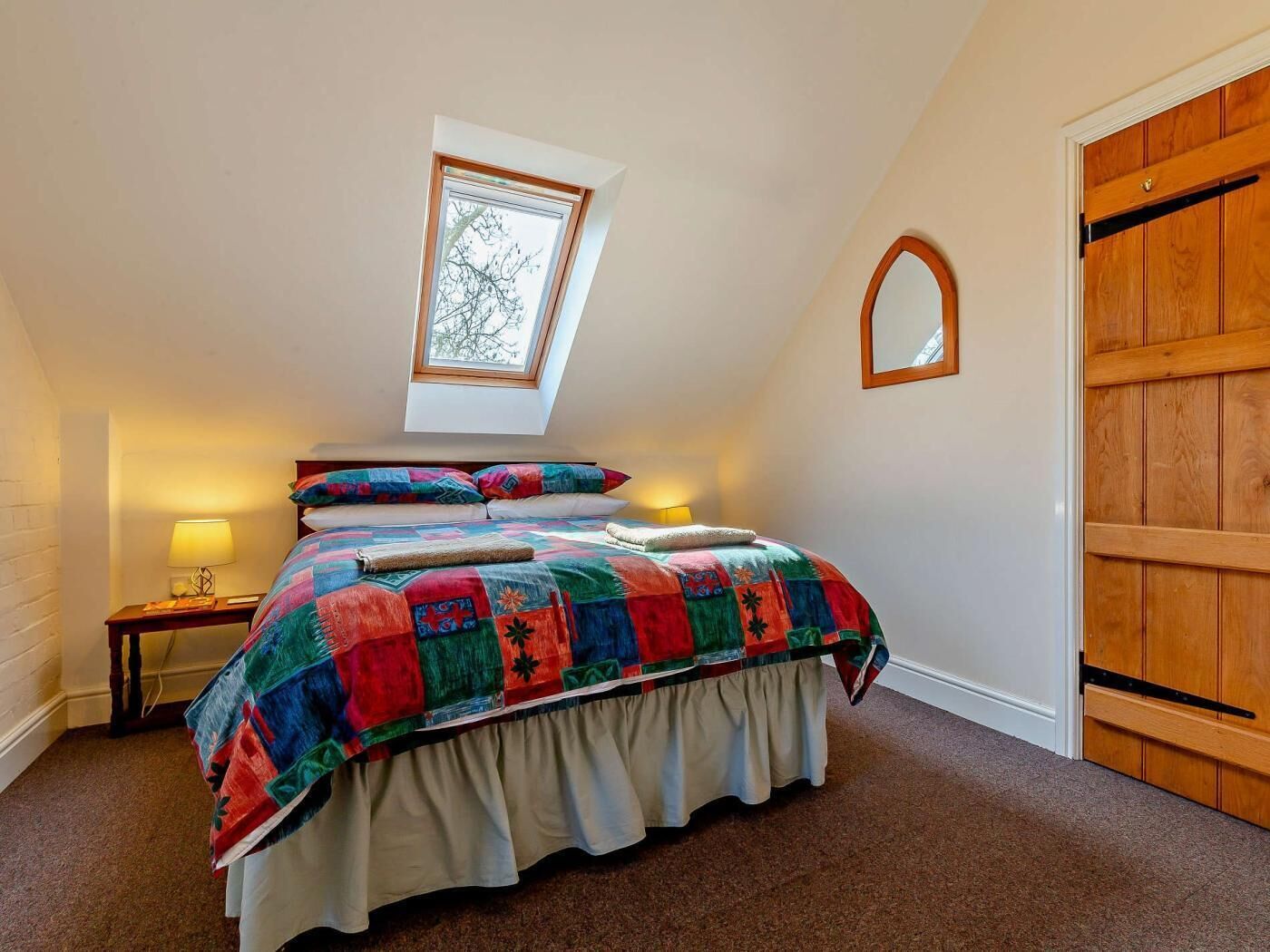 Une chambre avec un puits de lumière, un lit recouvert d’une courtepointe aux couleurs vives, une lampe de chevet et une porte en bois.