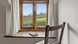 Et værelse med et vindue med træramme, hvide gardiner, en træstol og et lille bord med glas og en bog.