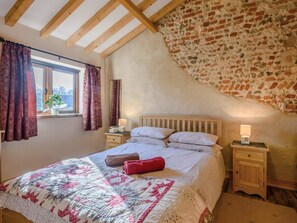 A bedroom with a wooden ceiling, a brick wall, a large bed with a quilt, two bedside tables with lamps, and a window with curtains. - 3 Bed in Corpusty (oc-ottn8) (Corpusty)