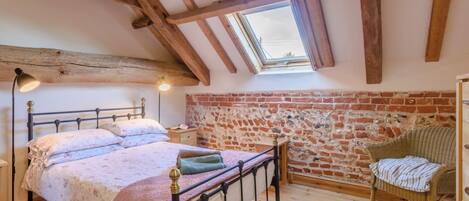 A bedroom with a sloped ceiling, exposed wooden beams, a brick wall, a bed with a headboard, a nightstand, a wicker chair, and a skylight.