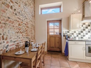 A kitchen with a stone wall, a wooden dining table with plates and cups, a window, and a tiled backsplash. - 3 Bed in Corpusty (oc-ottn8) (Corpusty)
