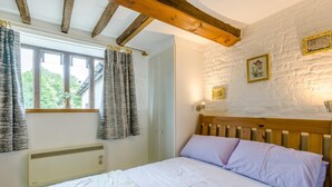 Une chambre à coucher avec une tête de lit en bois, un lit recouvert d’une literie blanche et lavande, une fenêtre avec des rideaux, un radiateur fixé au mur et un mur blanc rustique orné de tableaux encadrés.