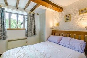 A bedroom with a wooden headboard, a bed with white and lavender bedding, a window with curtains, a wall-mounted heater, and a rustic white wall with framed pictures. - 1 Bed in Norwich (oc-stbn8) (Mulbarton)