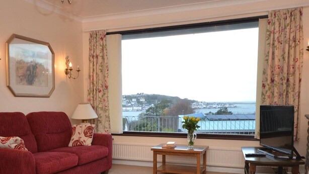 A living room with a red sofa, a small wooden table, a TV, and a view of a coastal landscape. - 2 Bed in Bideford (oc-sandb) (Instow)