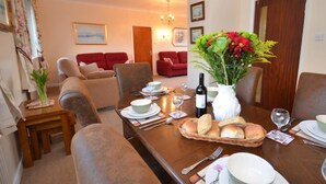 A dining area with a wooden table set for a meal, surrounded by chairs. There are two sofas in the background, a vase with flowers on the table, and a bottle of wine. - 2 Bed in Bideford (oc-sandb) (Instow)