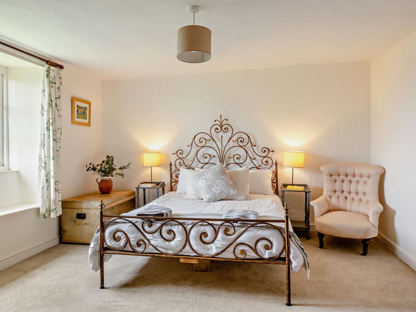 A bedroom with a metal bed frame, two bedside lamps, a chair, a window with curtains, and a small table with a plant.