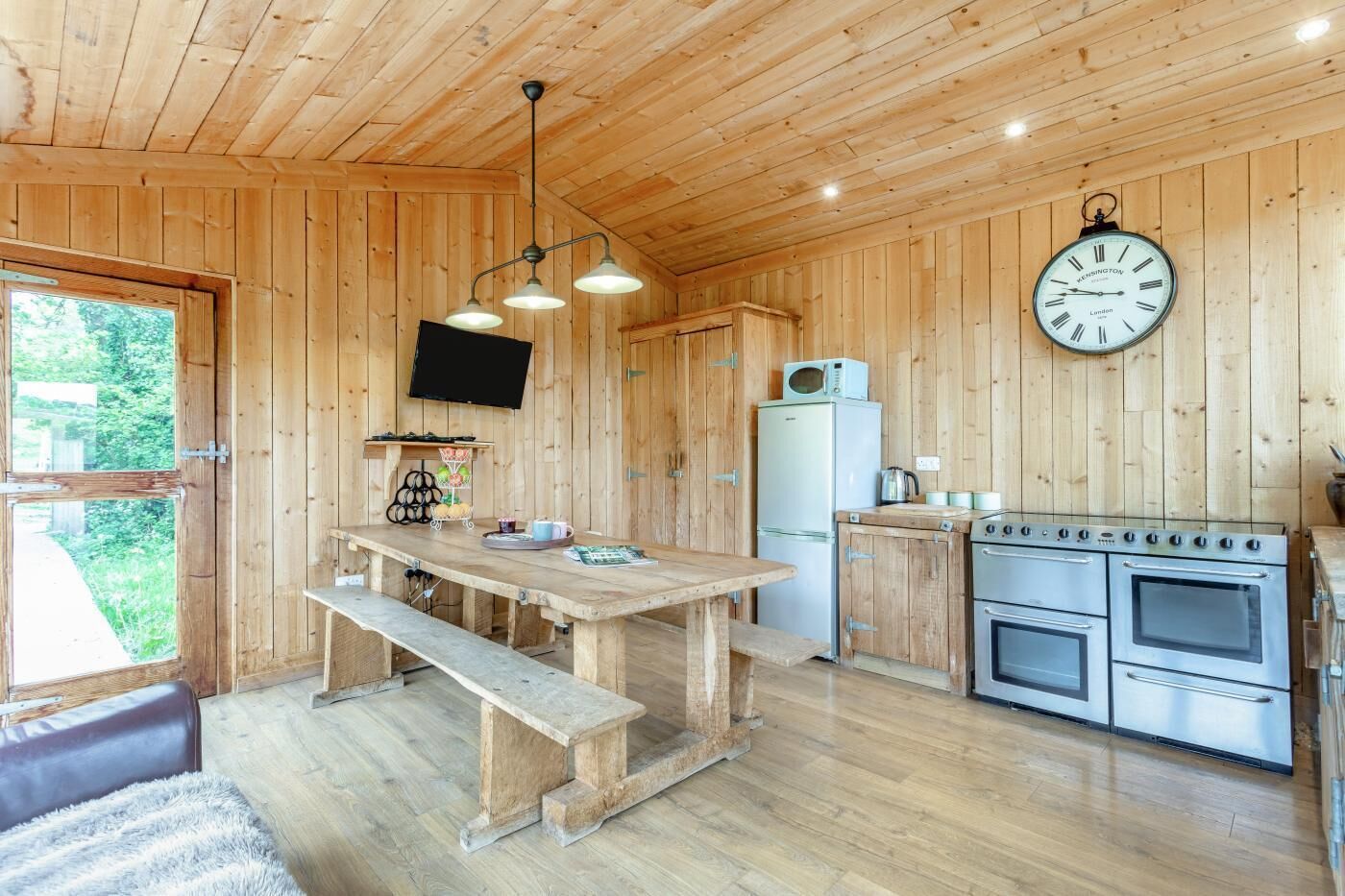 Ein Holzblockhaus-Interieur mit Holztisch, Bank, Küchenbereich inklusive Herd und Kühlschrank, sowie einer Wanduhr.