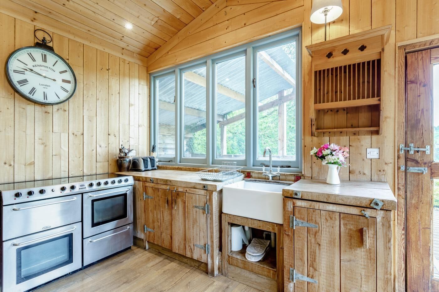A wooden kitchen with a large clock, stainless steel appliances, and a window with a view of greenery.