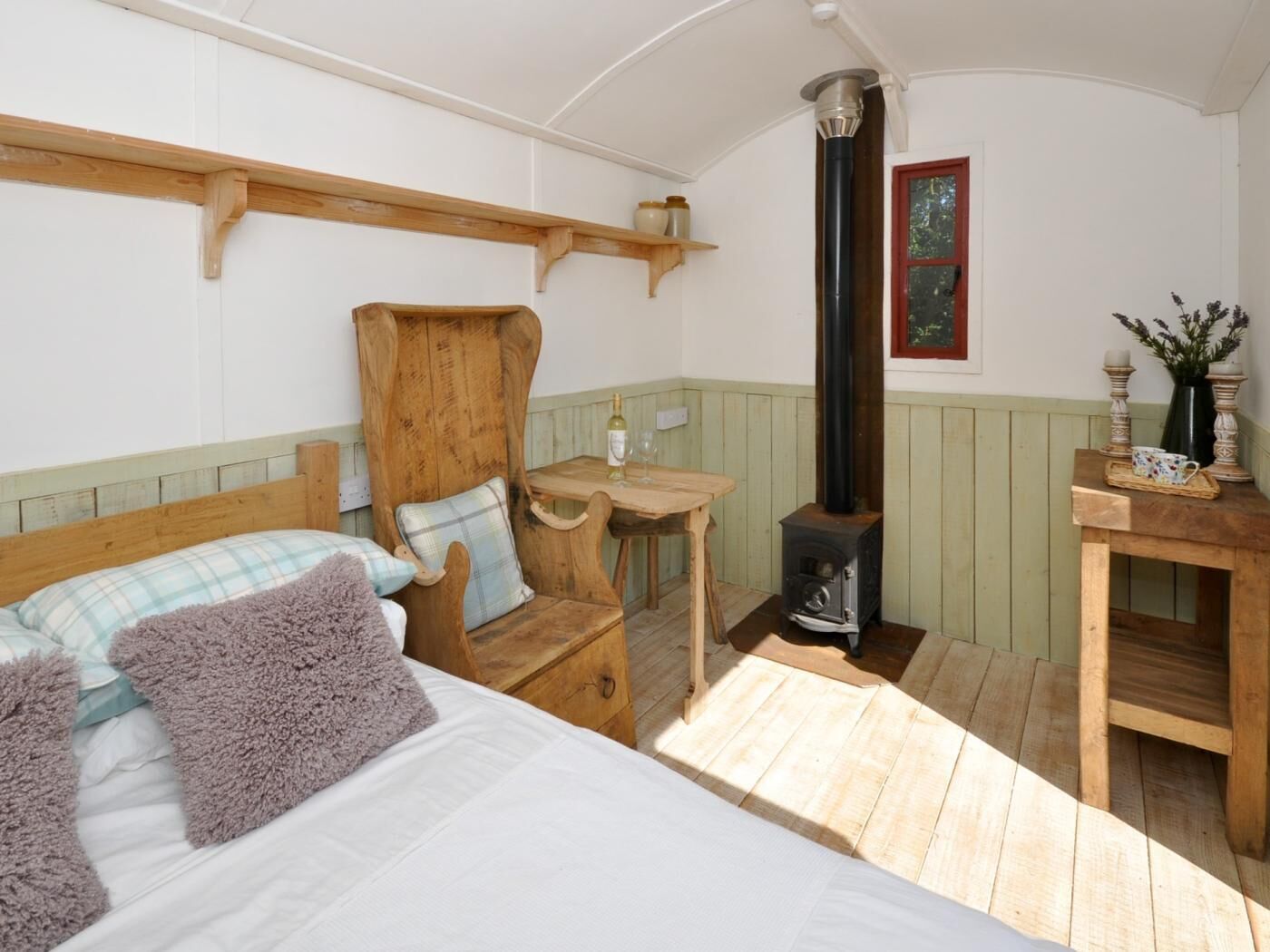 A cozy room with a bed, a wooden chair, a small table, a wall-mounted shelf, a fireplace, and a window with a view.