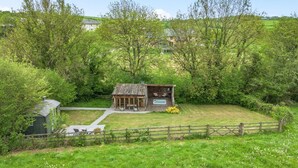 A rustic wooden cabin with a porch, surrounded by lush greenery and a fenced yard. - 1 Bed in Exeter (oc-lchas) (Cadbury)