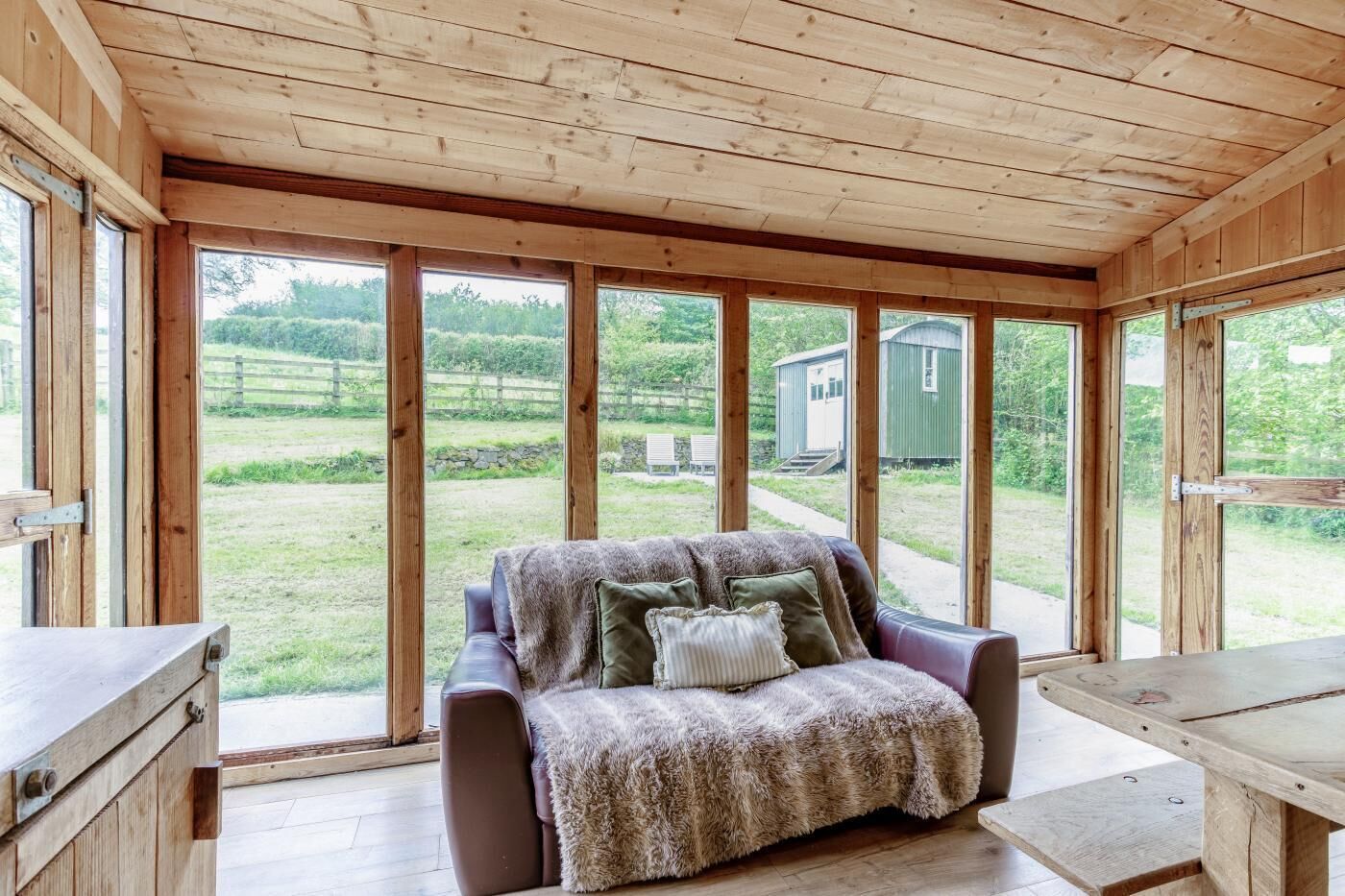 A cozy room with a sofa, a table, and large windows offering a view of a green field and a small shed.