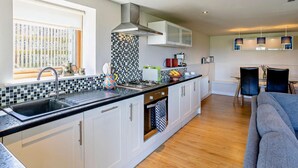 A modern kitchen with a large island, white cabinetry, a black countertop, and a window with blinds. - 2 Bed in Holmfirth (oc-emley) (New Mill)