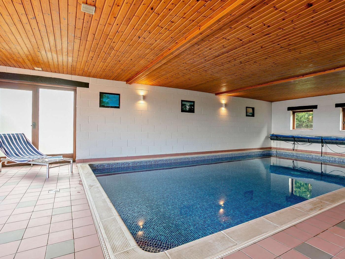 Ein beheiztes Hallenbad mit gefliesten Wänden, Holzbalkendecke und einer Liege.