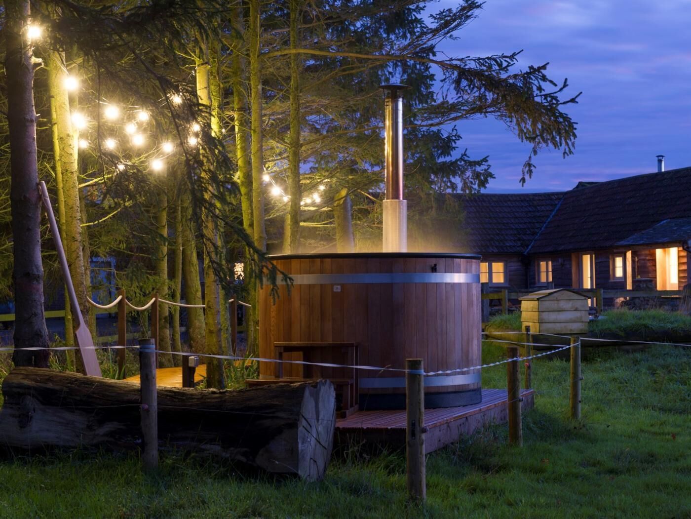 Een houten sauna met een metalen schoorsteen, omgeven door een houten terras en een touwhal, gelegen in een bebost gebied met een huis op de achtergrond.
