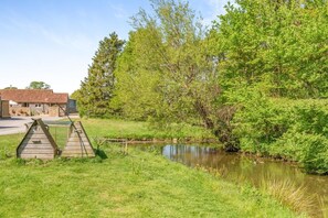 A rustic wooden structure near a pond, surrounded by lush greenery and a stone building in the background. - 3 Bed in Sturminster Newton (oc-cowob) (Hazelbury Bryan)