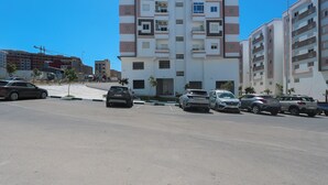 A parking lot with several cars, a multi-story residential building with balconies, and a clear blue sky. - TGV 7 min Corniche Plage WiFi fibre (Tanger)