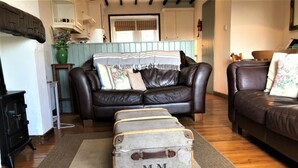 Un salon avec un canapé en cuir marron, une valise ancienne portant l’inscription « M.M. », et un plancher en bois.