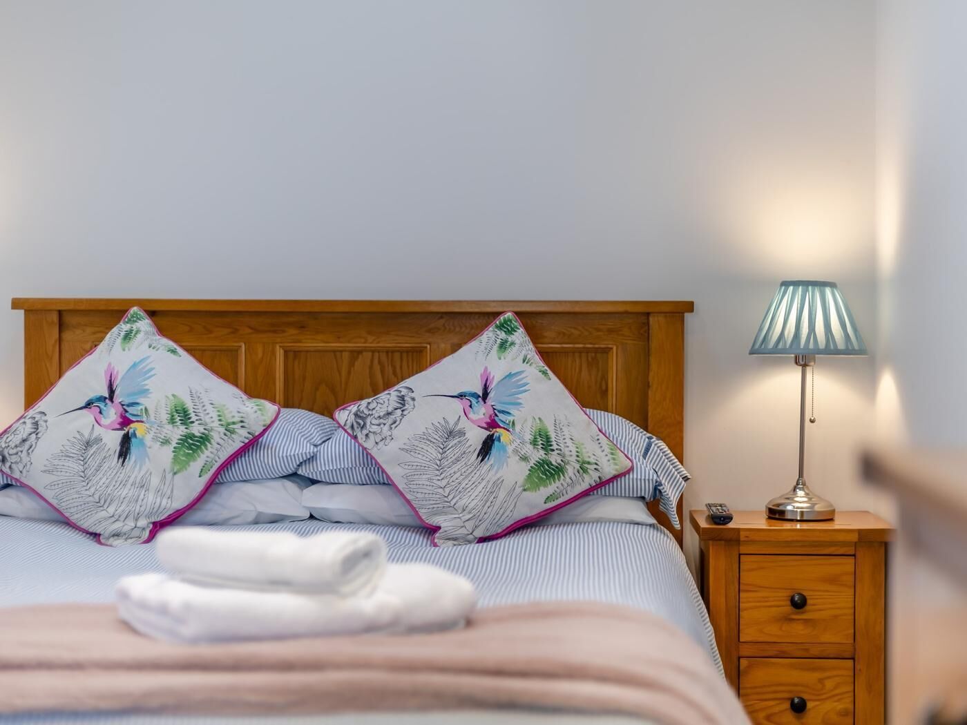 Una cama bien tendida con ropa de cama a rayas y dos almohadas decorativas con motivos de hojas y pájaros. También se observa una lámpara de noche y una mesita de noche de madera con un cajón.