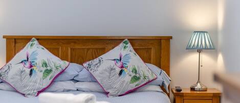 Una cama bien tendida con ropa de cama a rayas y dos almohadas decorativas con motivos de hojas y pájaros. También se observa una lámpara de noche y una mesita de noche de madera con un cajón.