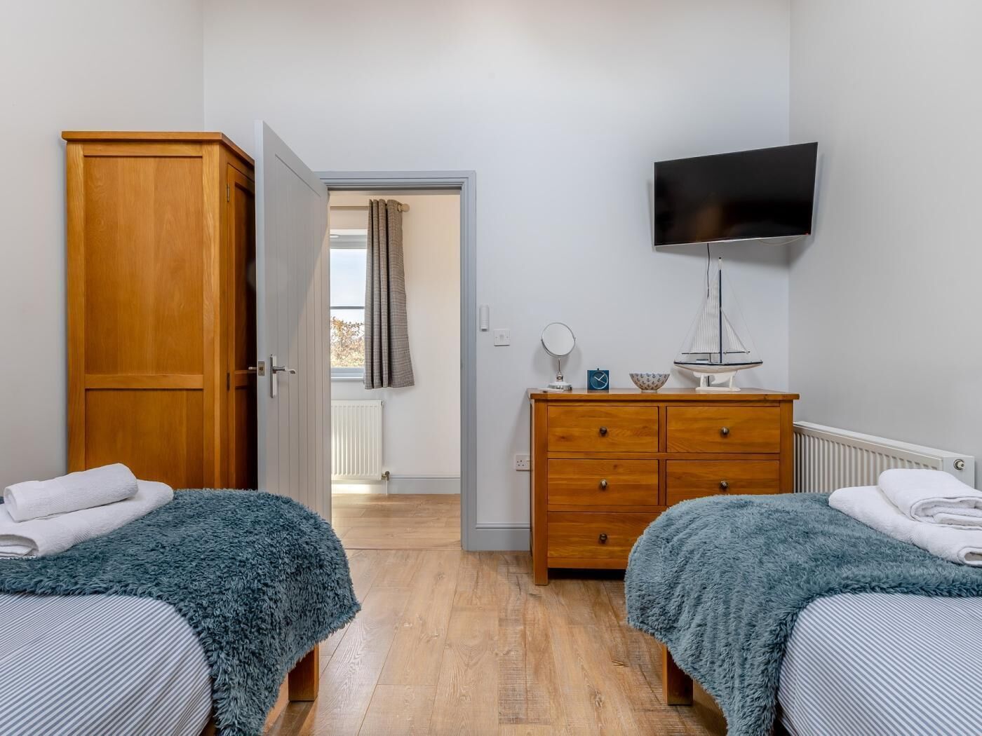 Ein Schlafzimmer mit zwei Betten, einem Holzschrank, einem Schminktisch und einem Flachbildfernseher. Ein Fenster mit Vorhängen ist ebenfalls vorhanden.