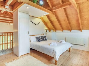 Et soveværelse med skrå vægge, træbjælker, en seng med hvide sengetøj, en sengebordslampe og en radiator.