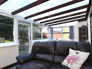 A sunlit room with a black leather sofa, a wooden cabinet, and a large window with a view of a backyard. - 1 Bed in Holsworthy (oc-wscot) (Pancrasweek)