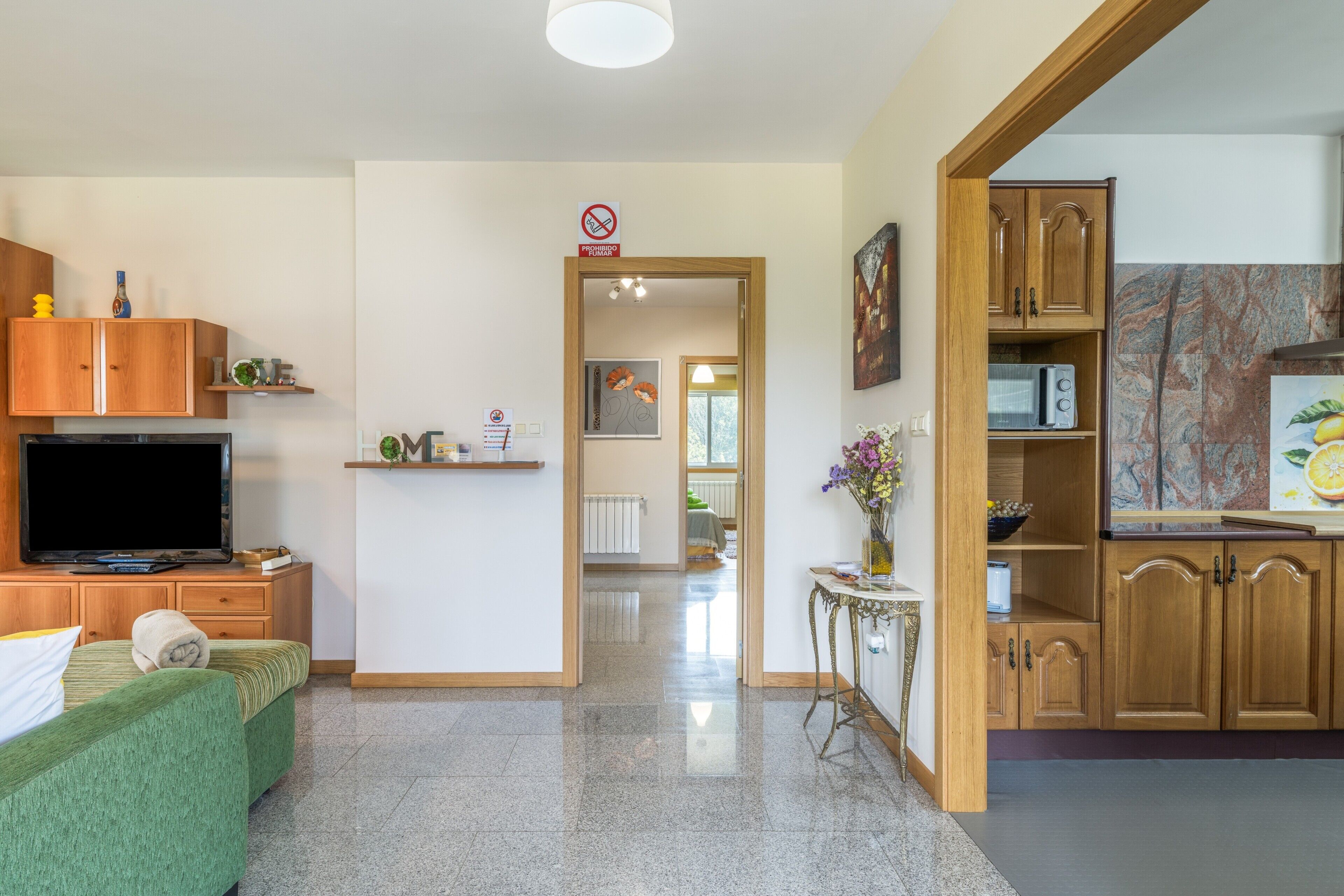 Habitación de hotel con sofá verde, armarios de madera, televisor, una mesita con un florero y una zona de cocina con armarios de madera y microondas.