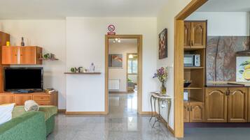 Habitación de hotel con sofá verde, armarios de madera, televisor, una mesita con un florero y una zona de cocina con armarios de madera y microondas.