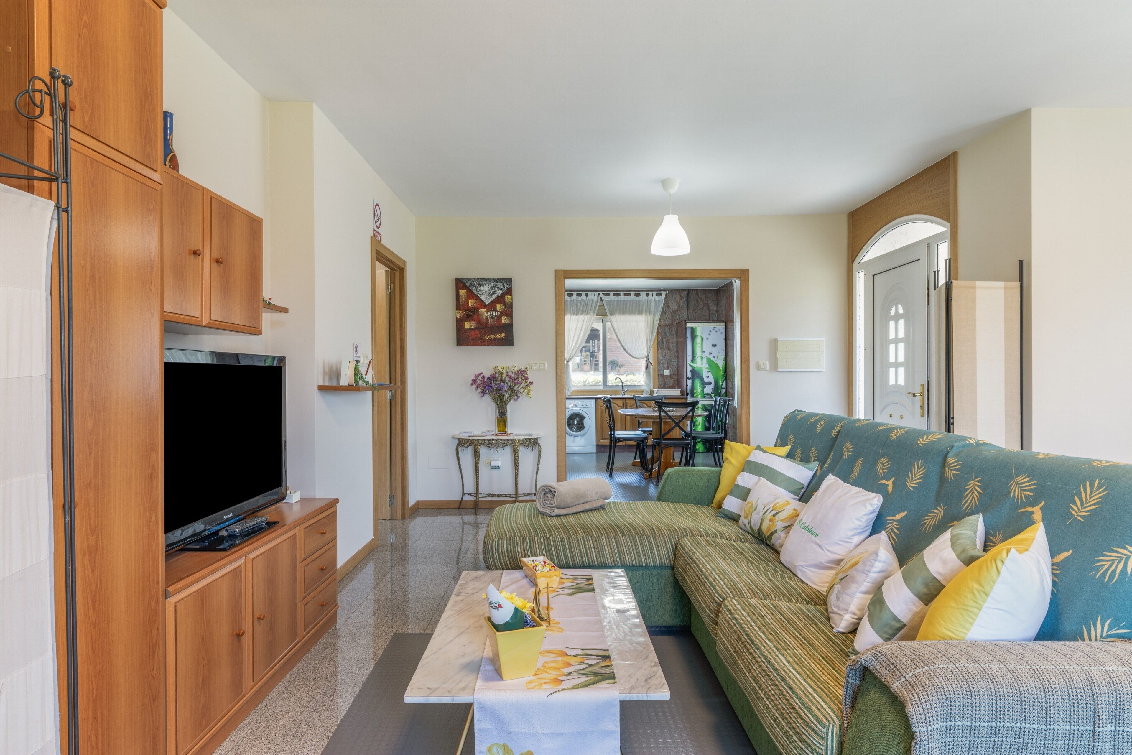 A living room with a sofa, television, wooden cabinets, and a dining area in the background.