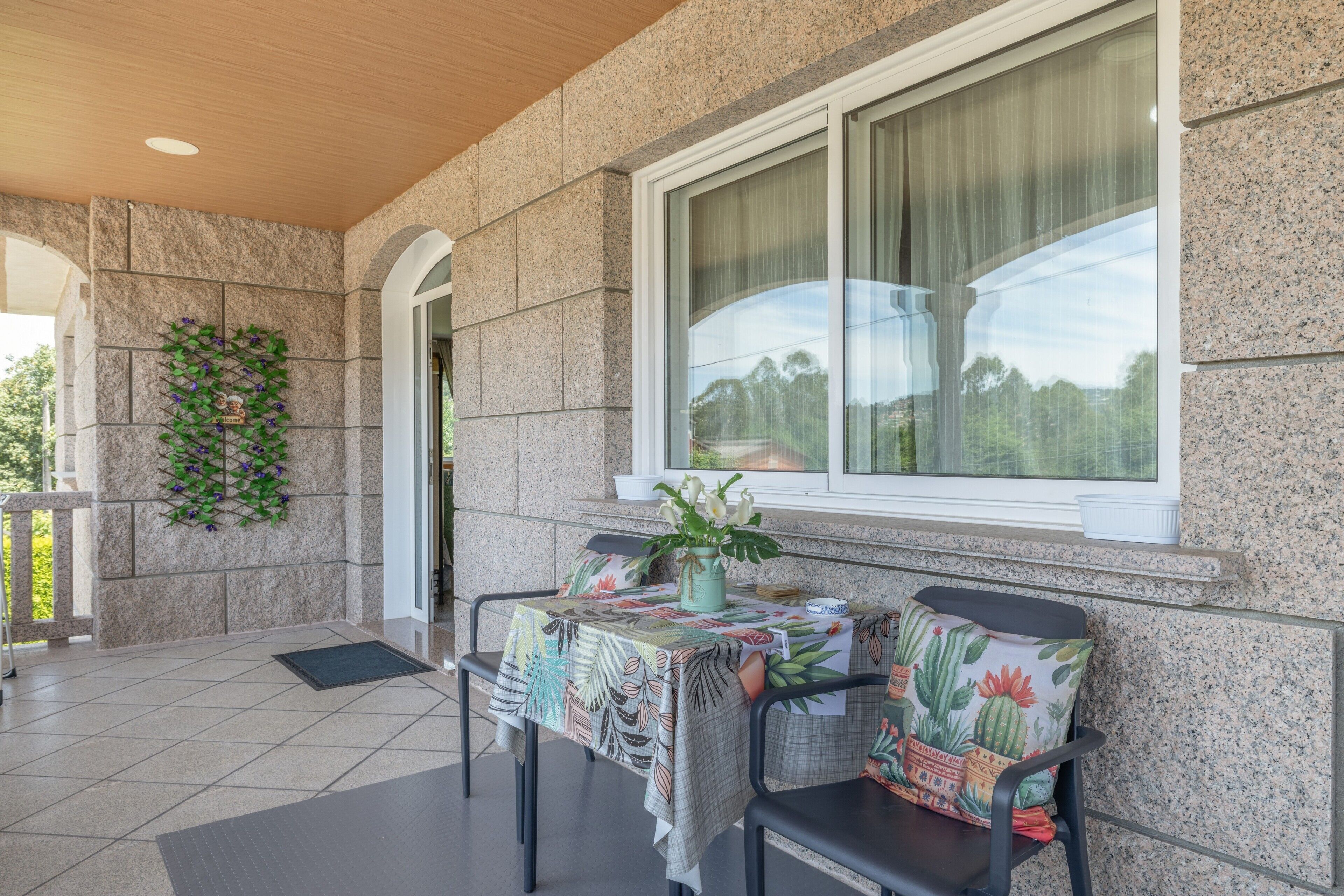 An outdoor seating area with a table covered in a floral tablecloth, two chairs with cactus-pattern cushions, and a window with curtains.