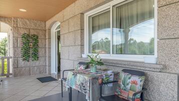 An outdoor seating area with a table covered in a floral tablecloth, two chairs with cactus-pattern cushions, and a window with curtains.