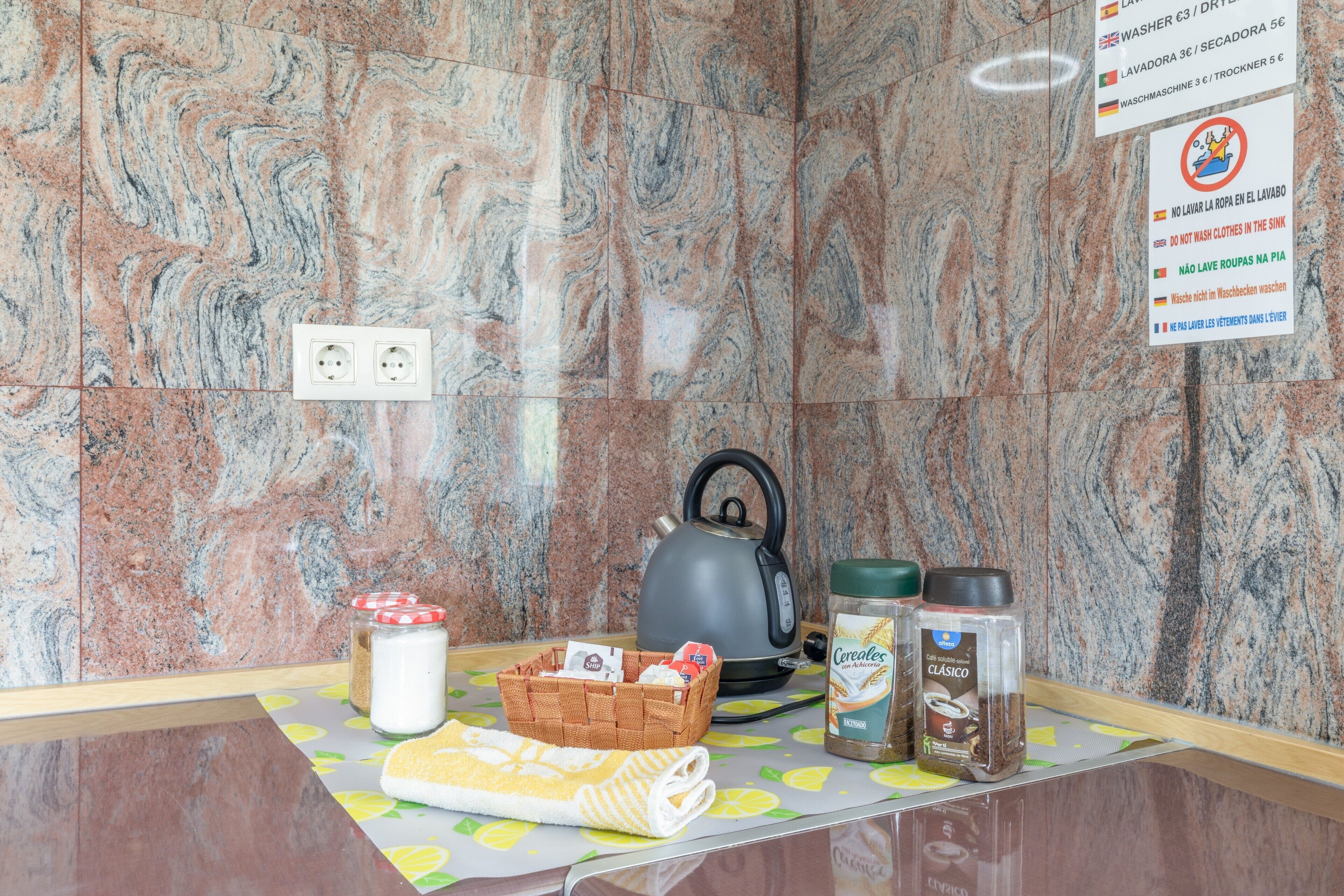 A kitchen counter with a kettle, jars, a basket, and a tablecloth.