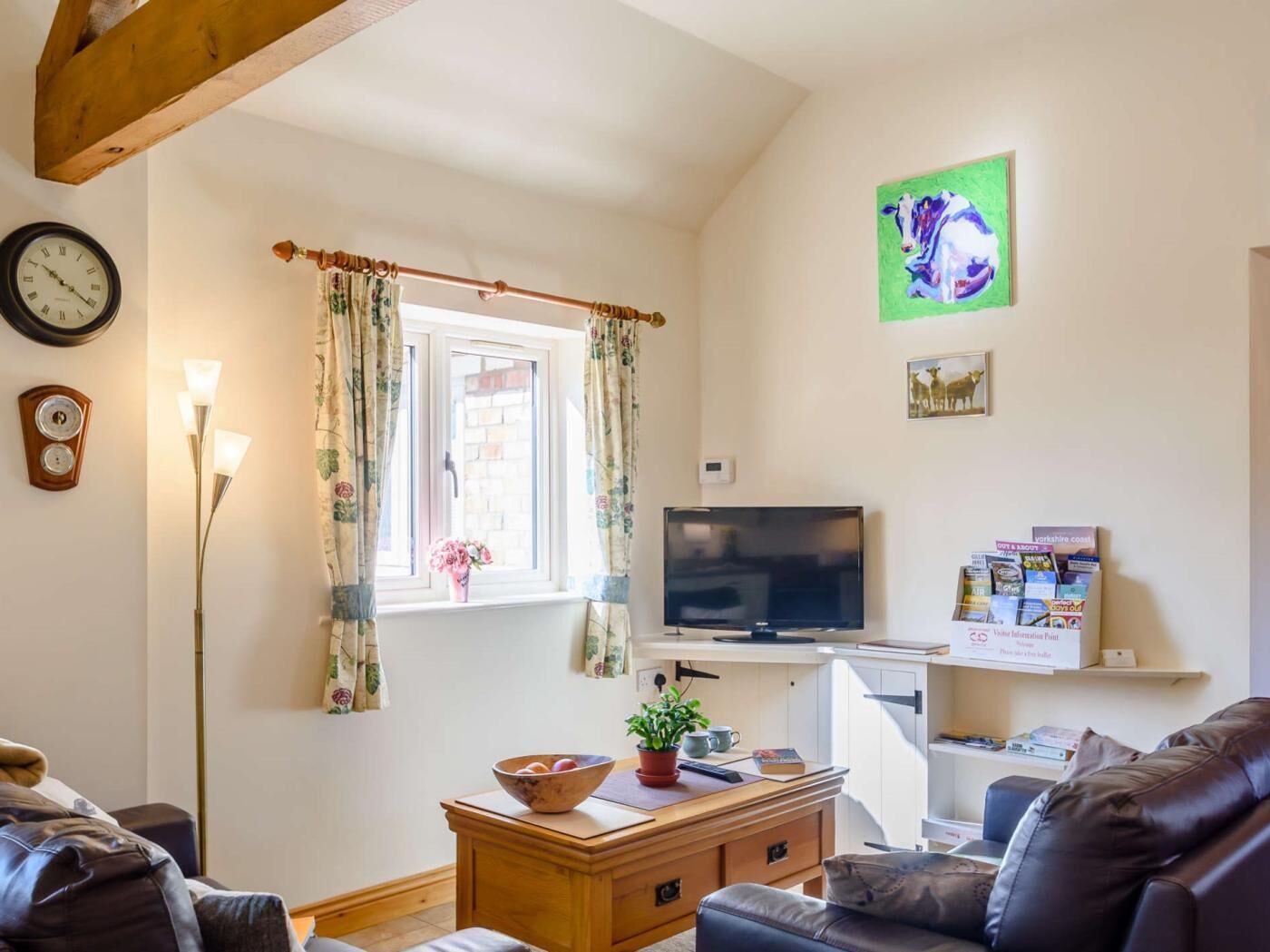 A living room with a television, a clock, a window with curtains, a plant, and a bookshelf.
