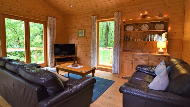 A wooden cabin interior with a TV, leather sofas, a wooden table, and a bookshelf.