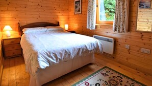 A wooden cabin room with a bed, nightstands, a window with curtains, and a radiator.