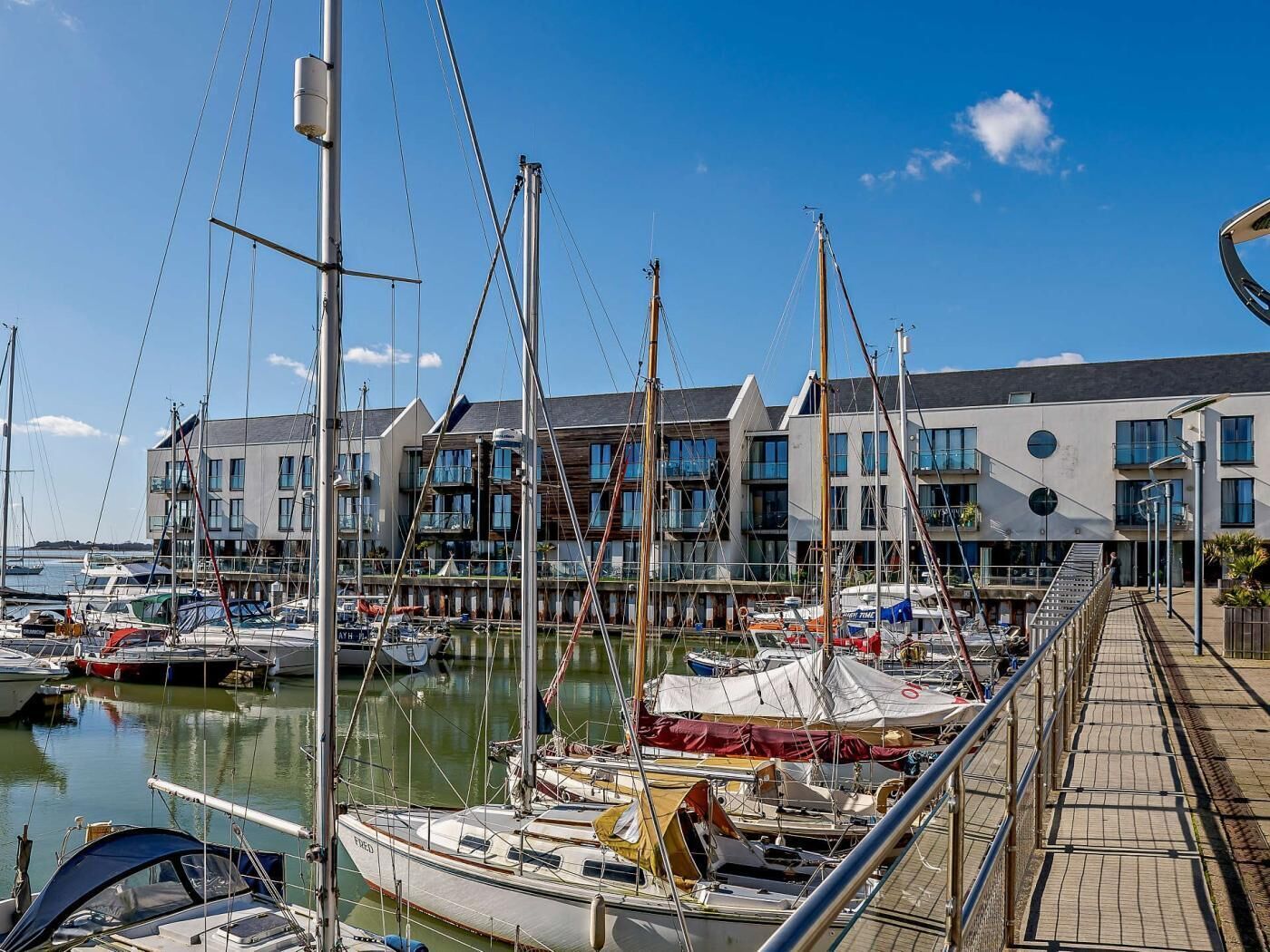 Un port de plaisance avec des bateaux amarrés, une promenade piétonne et des bâtiments modernes dotés de balcons.