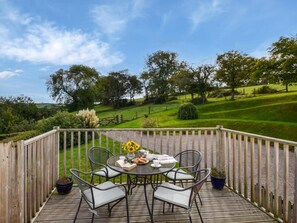 Una terraza de madera con una mesa y sillas, con vistas a un paisaje verde con árboles y colinas.