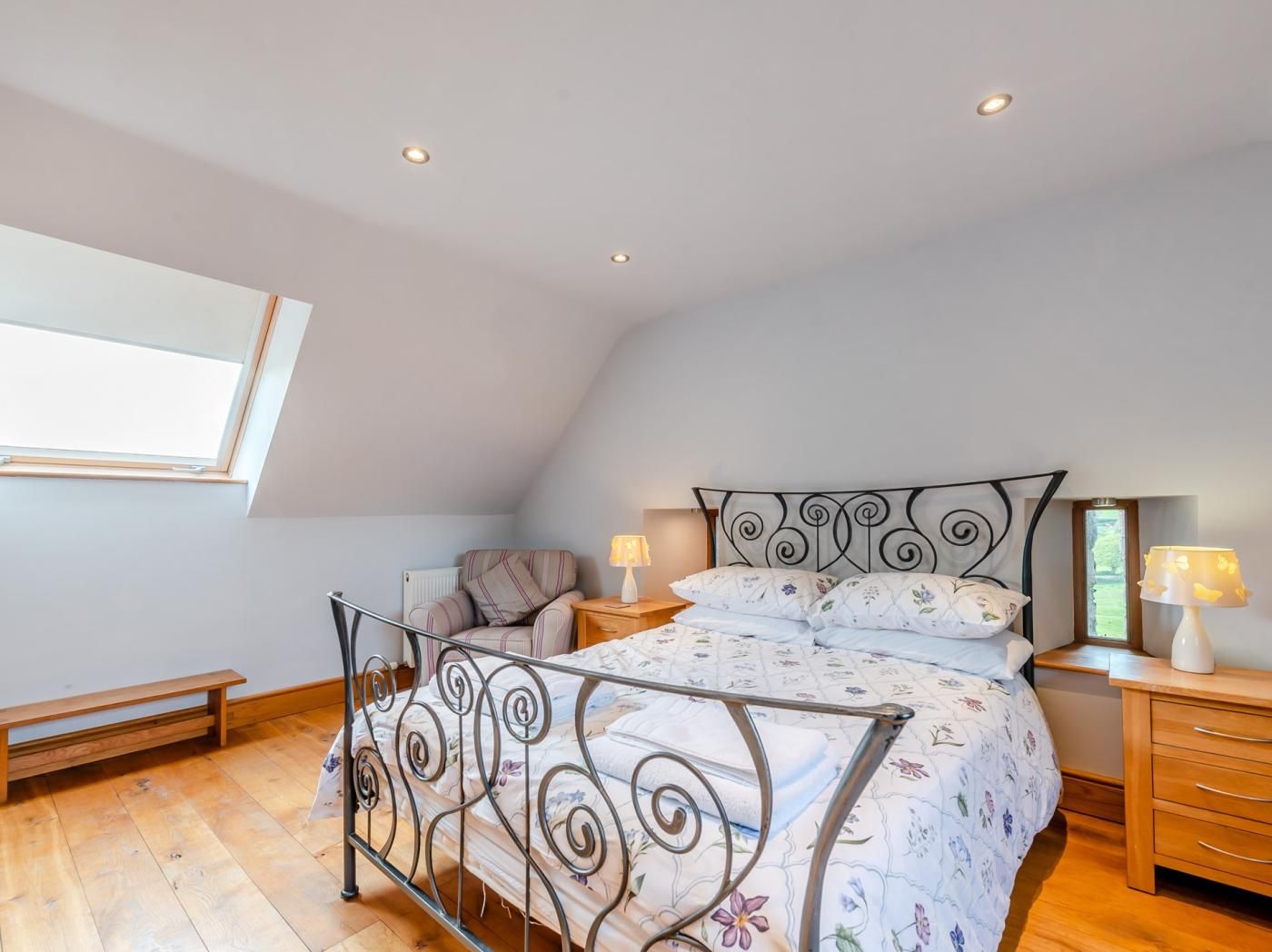 Une chambre avec un plafond en pente, un lit avec un cadre en métal, une table de chevet avec une lampe, un banc et une fenêtre.
