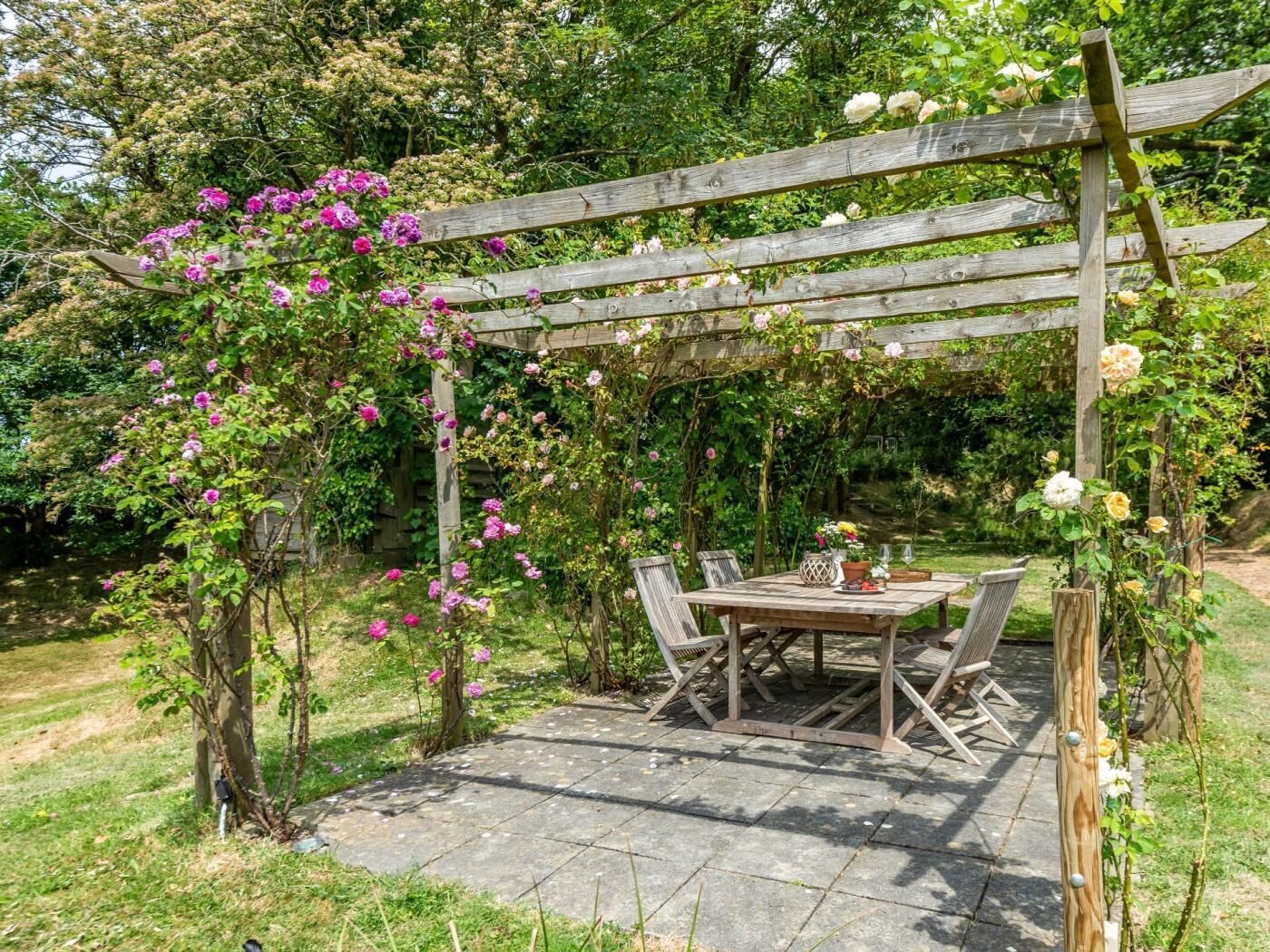 Une pergola en bois ornée de plantes grimpantes, une terrasse en pierre et une table avec des chaises.