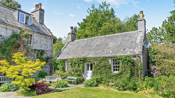 A stone house with a tiled roof, a garden with benches, and a clear sky.